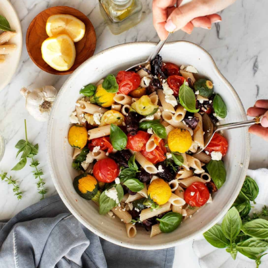 A bowl of Italian pesto pasta with roasted cherry tomatoes, zucchini, and red bell peppers.